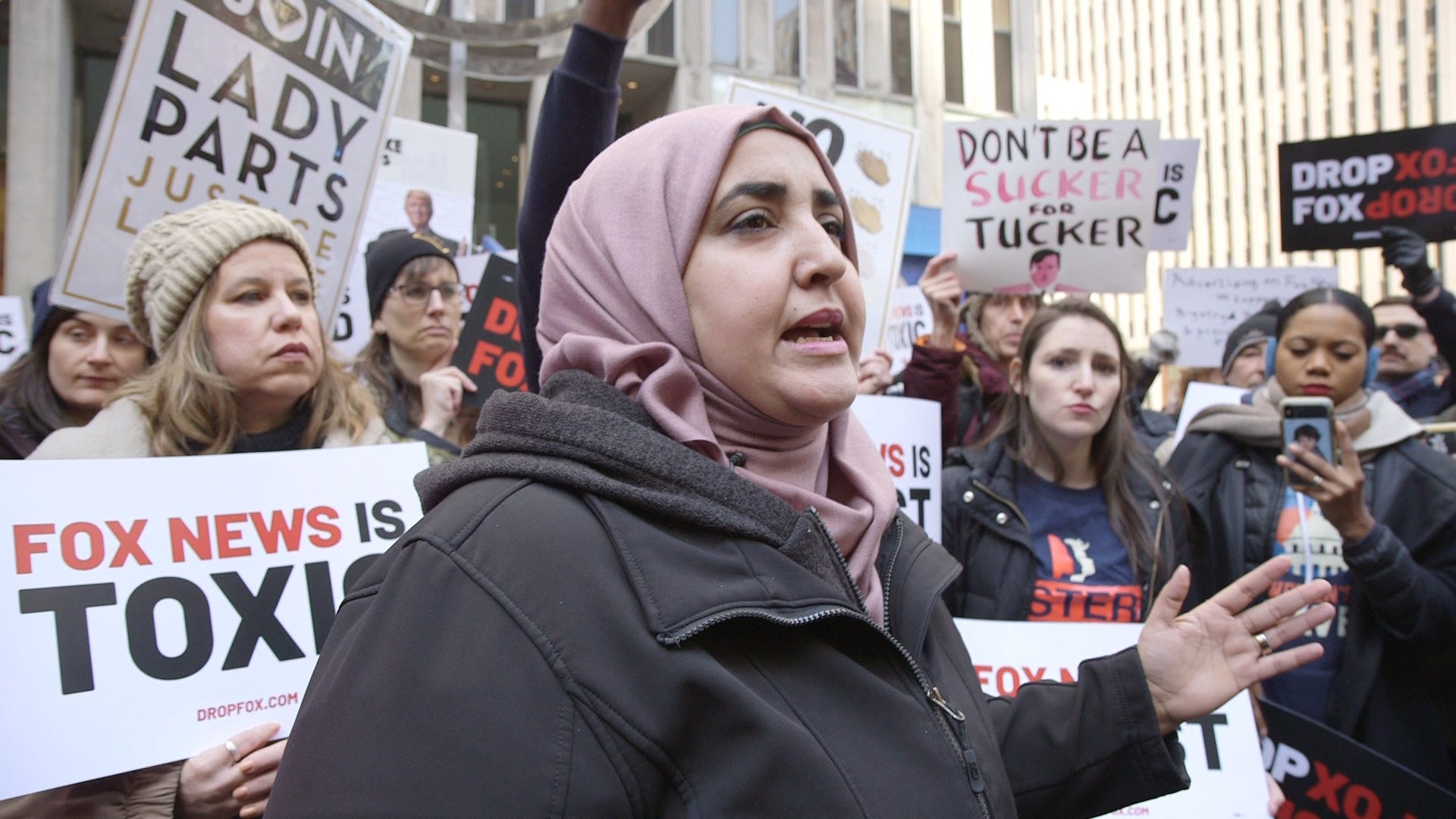 A woman in a hijab stands amongst a crowd of women holding signs during a protest or gathering.