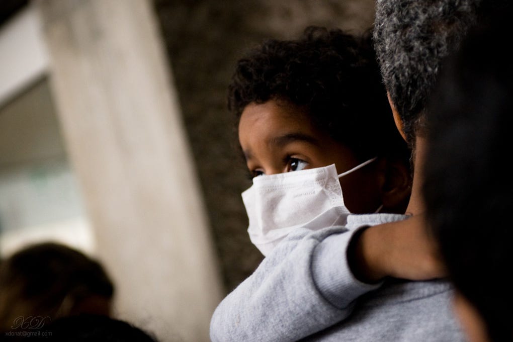 Small child with dark skin and curly black hair wears a face mask while being carried by an adult. The child looks over the adult’s shoulder, clinging to the adult, eyes wide.