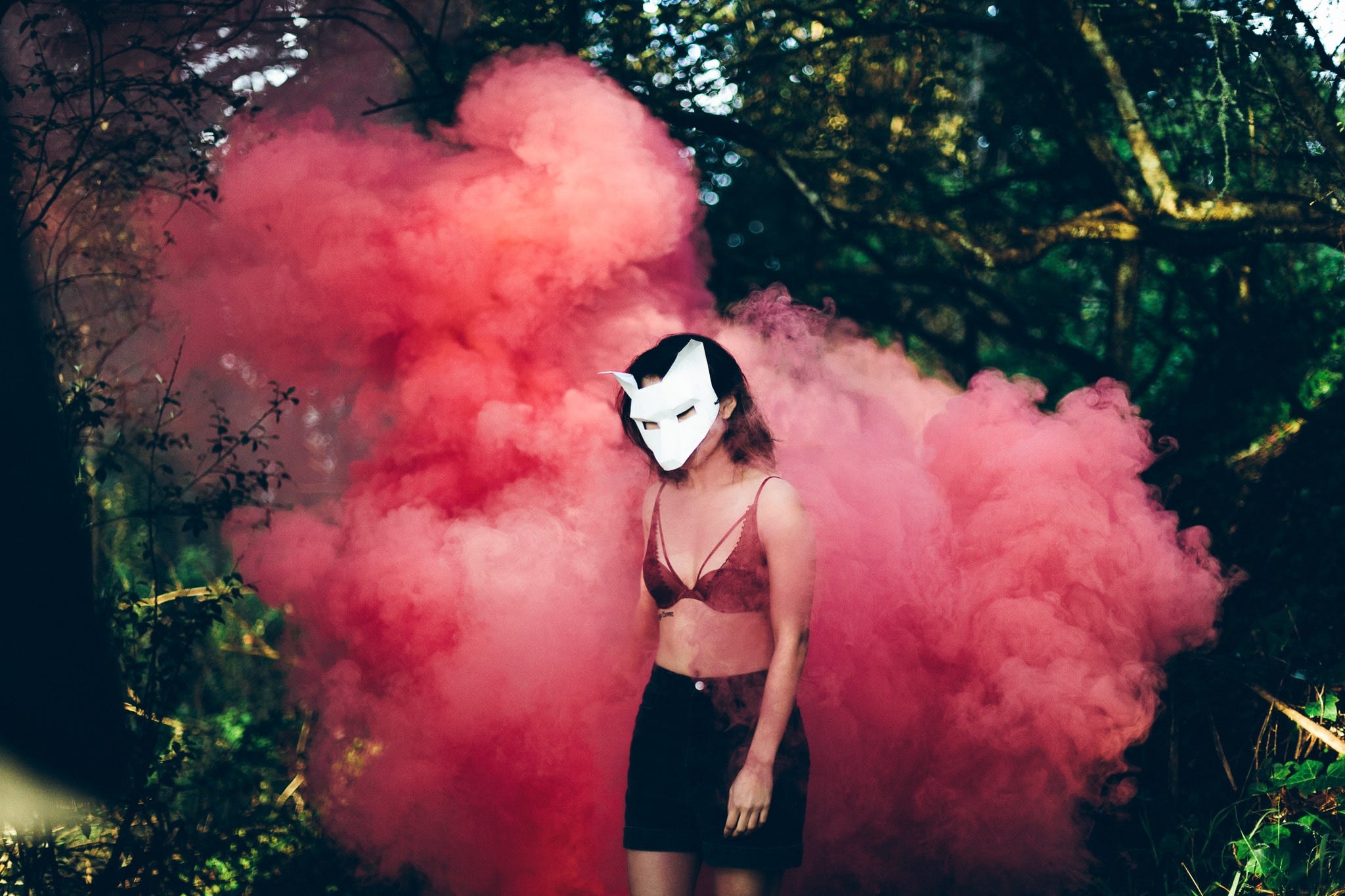 Person in the forest stands in front of a pink smoke cloud, wearing a paper wolf mask, a red bra and a black skirt.
