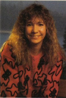 An older photograph. A young woman with wavy, shoulder-length hair and dark eyes smiles at the camera. She is wearing a pink shirt with black striped designs, and is against a blue-grey background.