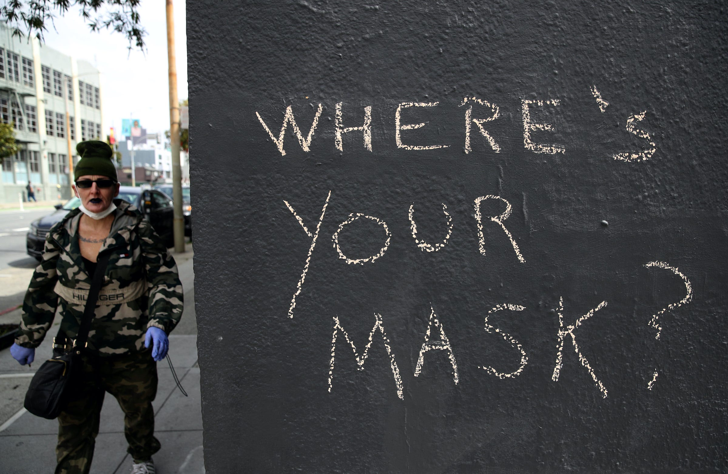 On the right side, three-quarters of the pic is a black exterior wall with “Where’s your mask?” written on it in chalk. The quarter of the photo on the left side is a person wearing latex gloves and a face mask, the latter under their chin.