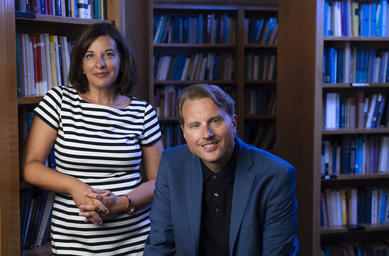 Annalisa Coliva stands next to a book case with Duncan Pritchard sitting next to her
