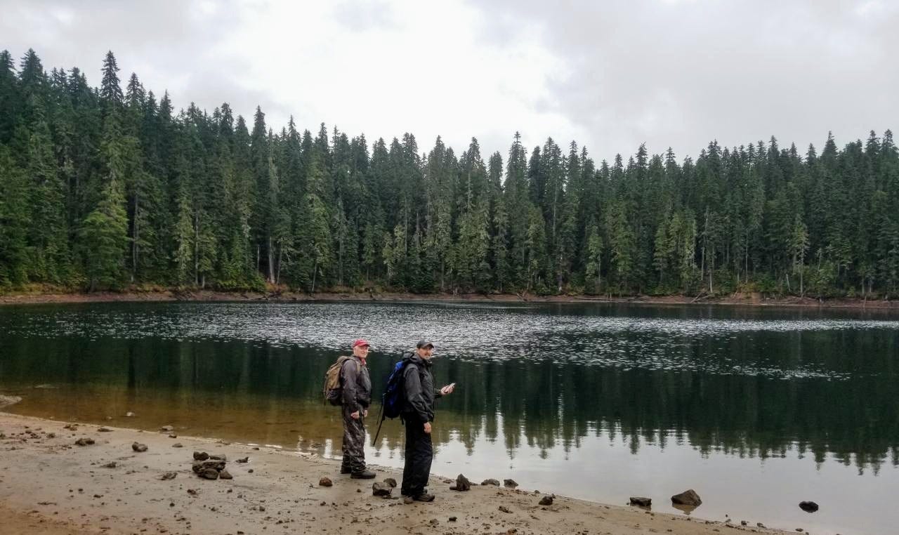 Two men stand at the waters edge of a lake with trees on the opposite side of the lake