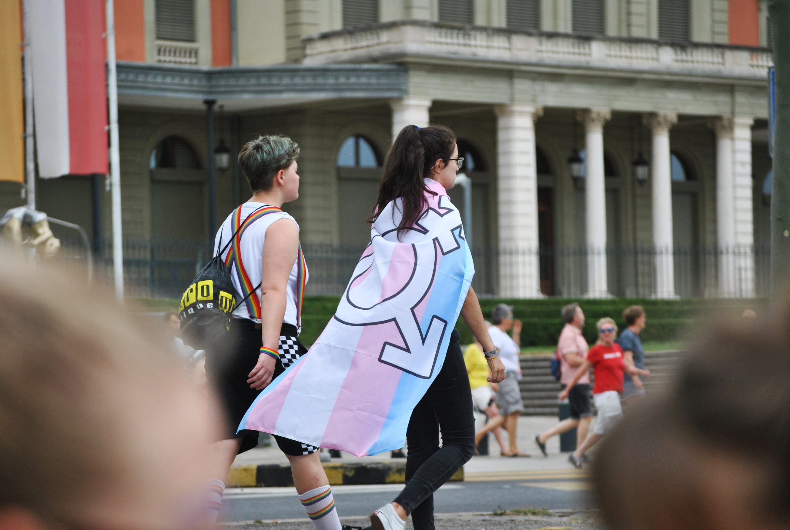 People marching down street, one wearing transgender flag as a cape