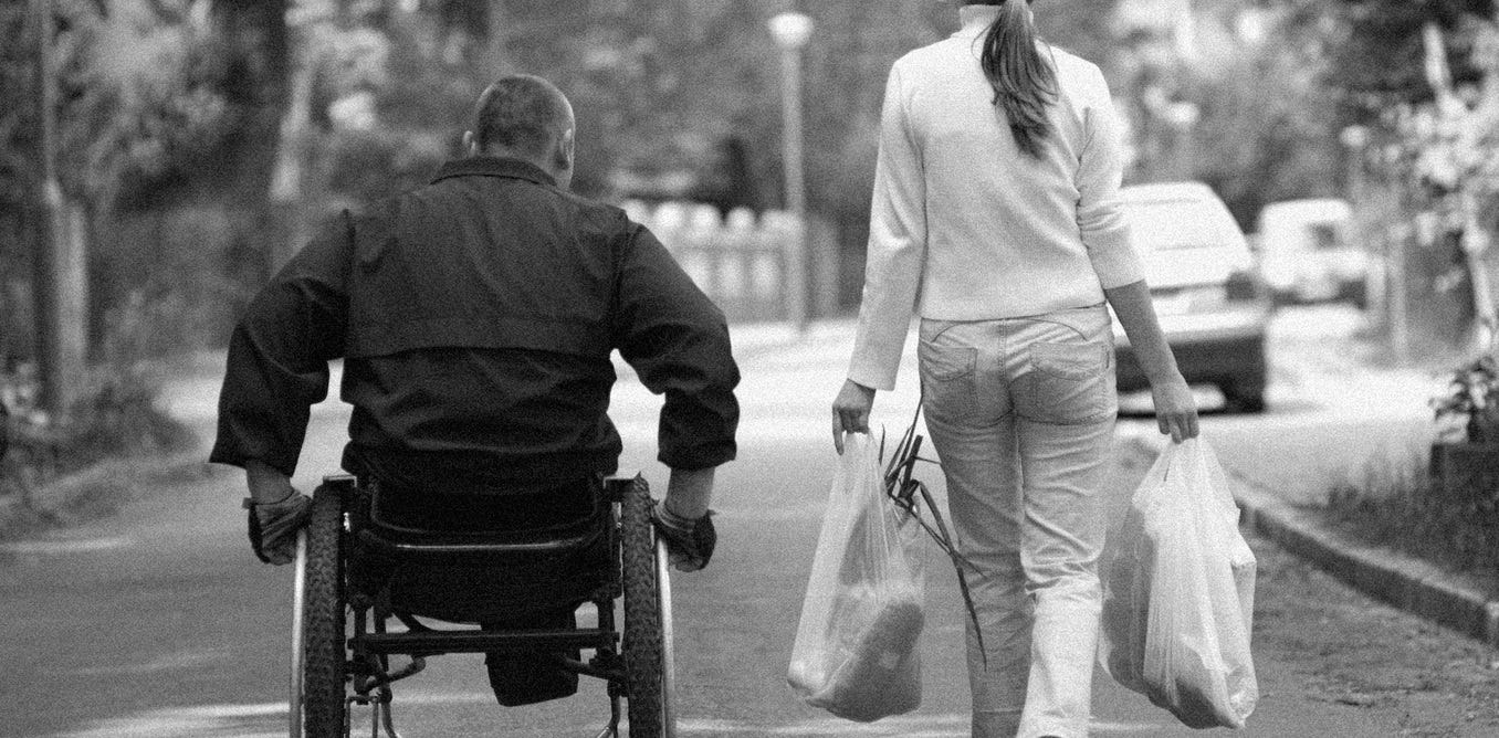 A man in a wheelchair next to a lady walking and carrying grocery bags with food.