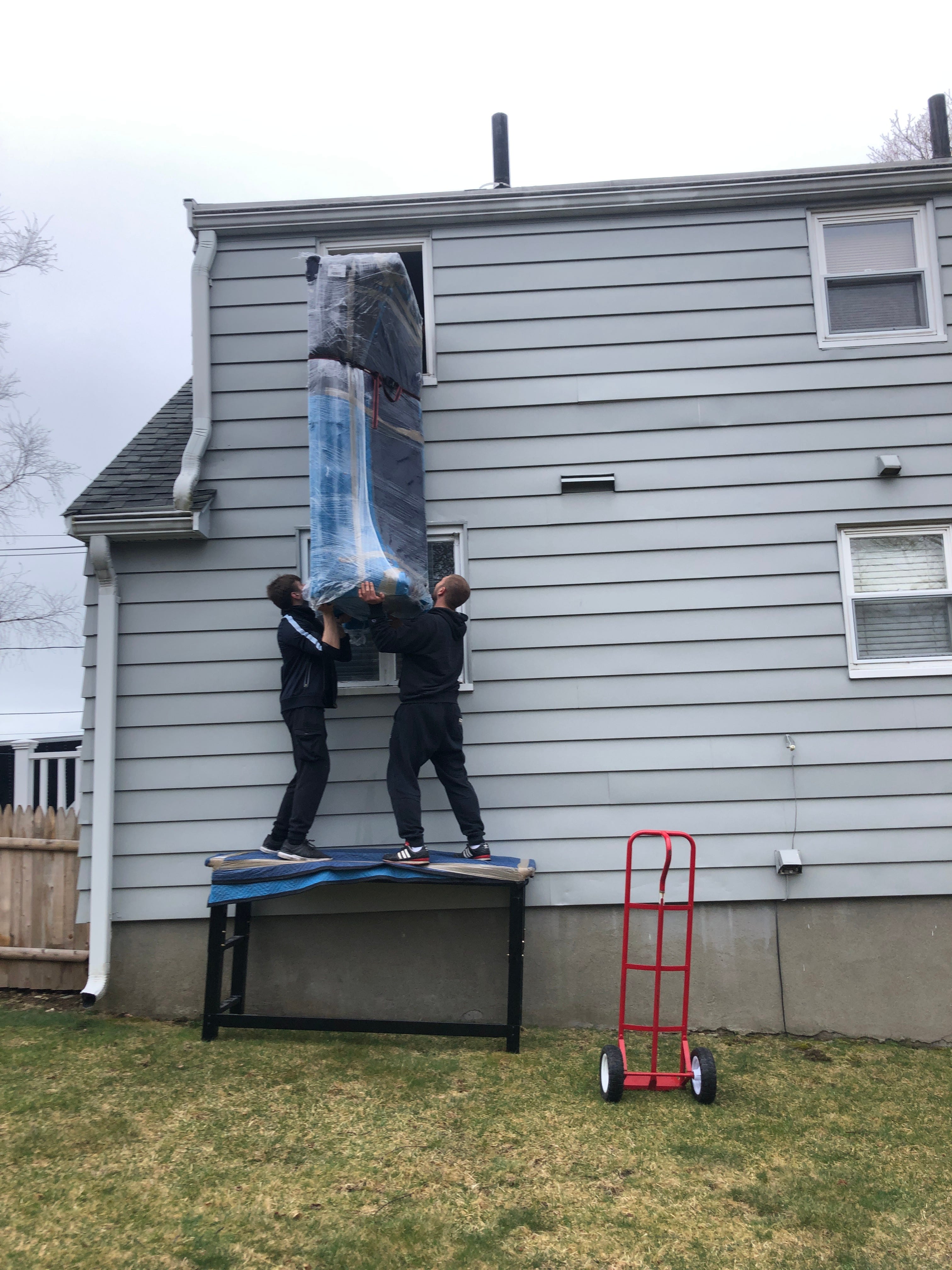 Two people standing on a table, outside, at the side of a house, hositing a sofa to a second-story window