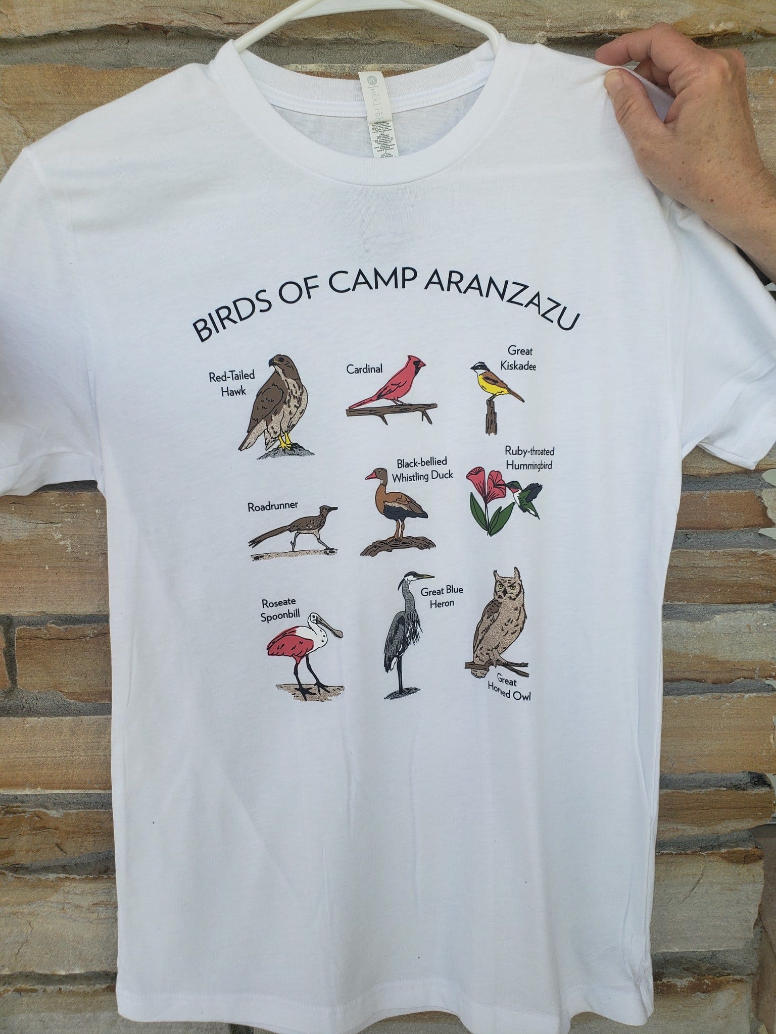 Photo ofCamp Aranzazu’s white birding t-shirt called Birds of Camp Aranzazu. The graphic includes caricatures of each of the following birds: Red-Tailed Hawk, Cardinal, Great Kiskadee, Roadrunner, Black-bellied Whistling Duck, Ruby-throated Hummingbird, Roaseate Spoonbill, Great Blue Heron, and the Great Horned Owl.
