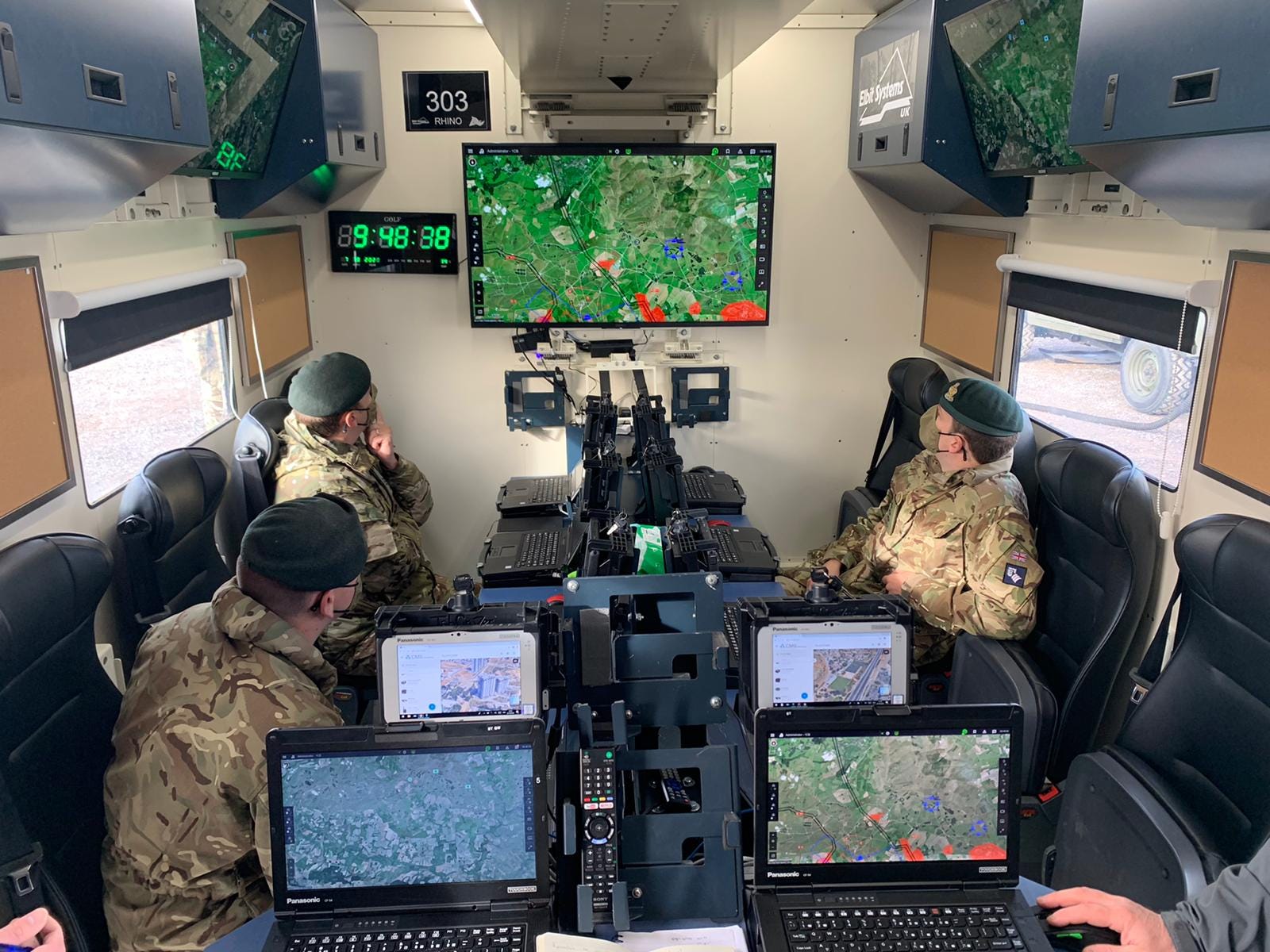 Three soldiers watching a big screen and smaller screens that is show an aerial view of a simulated battlefield.