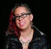 Queer non-binary Autistic femme of Burgher/Tamil Sri Lankan and Irish/Roma ascent, with curly dyed red hair, wearing glasses and smiling.
