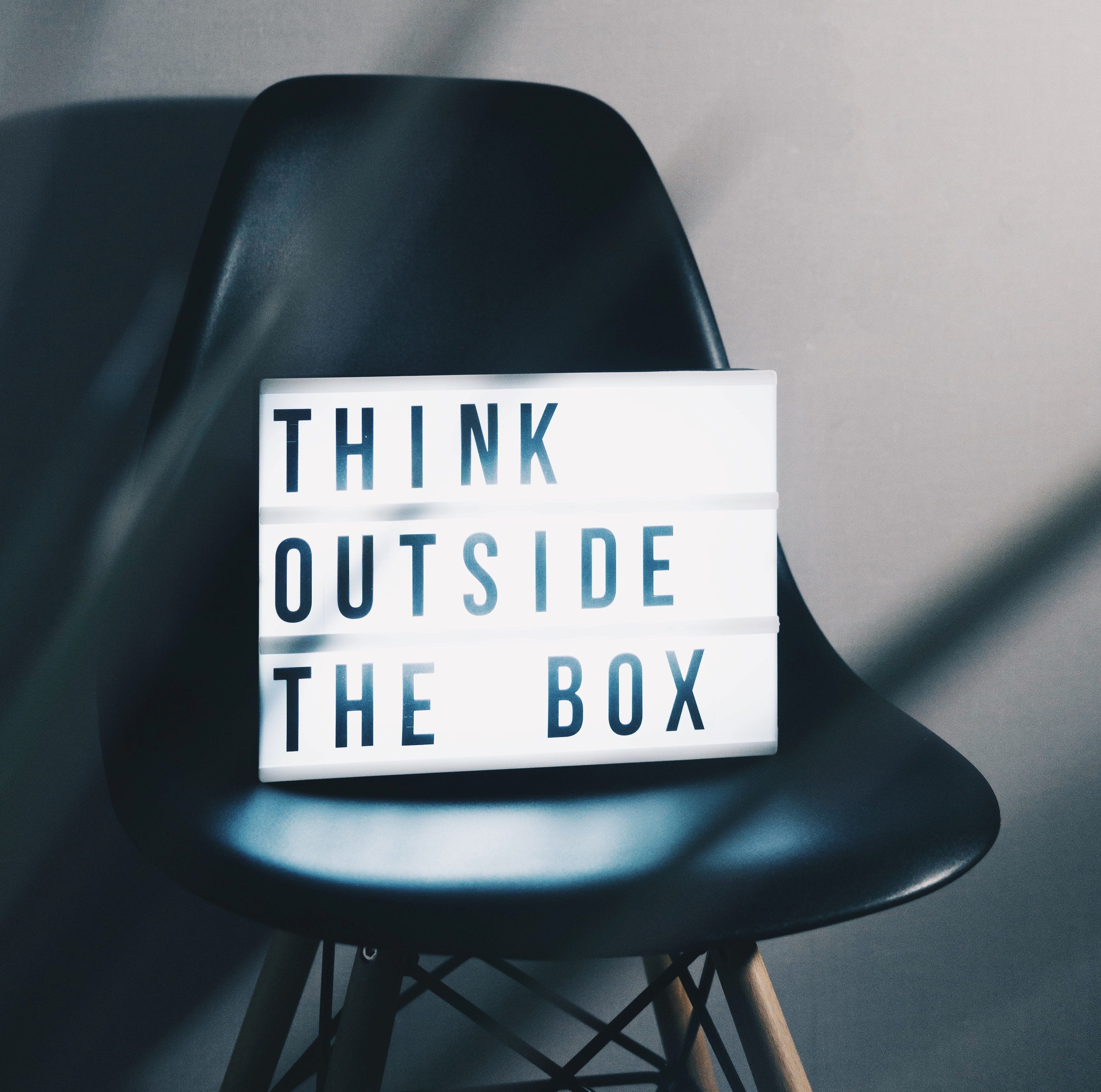 A white light box sign with black letters saying ‘Think outside the box’. The sign is sitting on black leather desk chair.