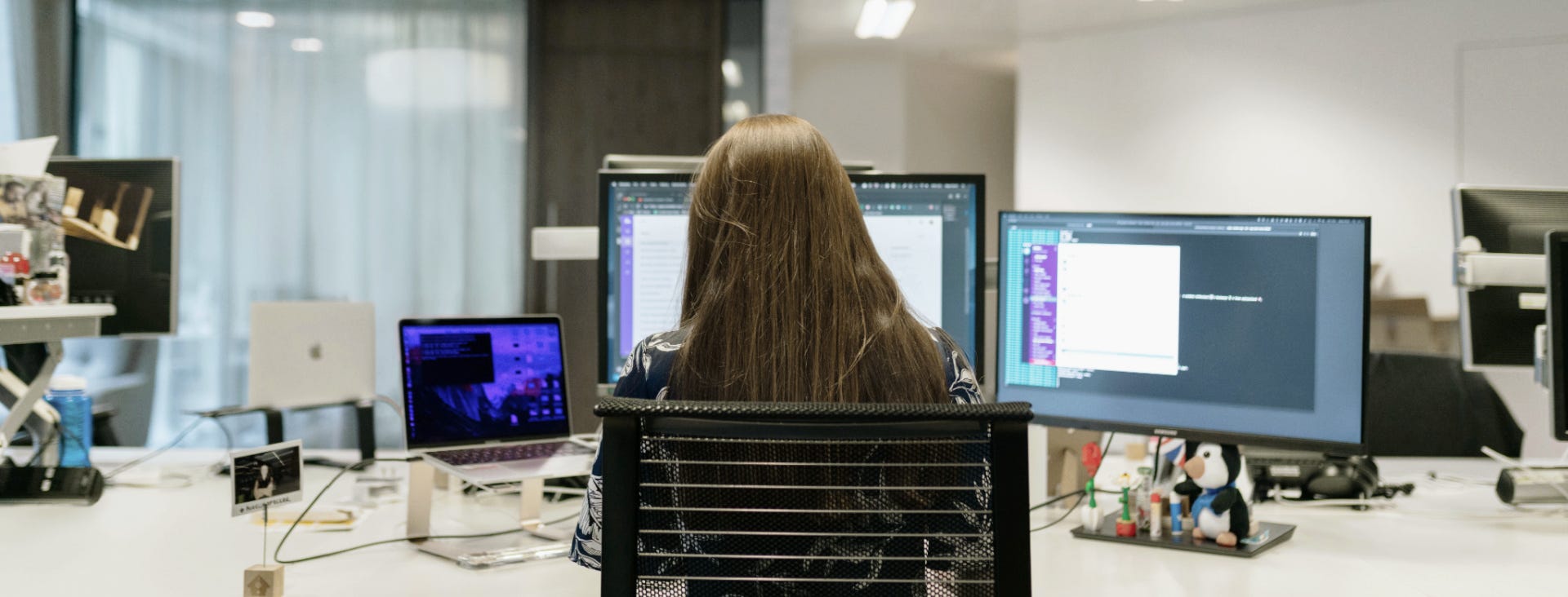 Back of a software engineer’s head as she reviews code on triple monitors in an office setting