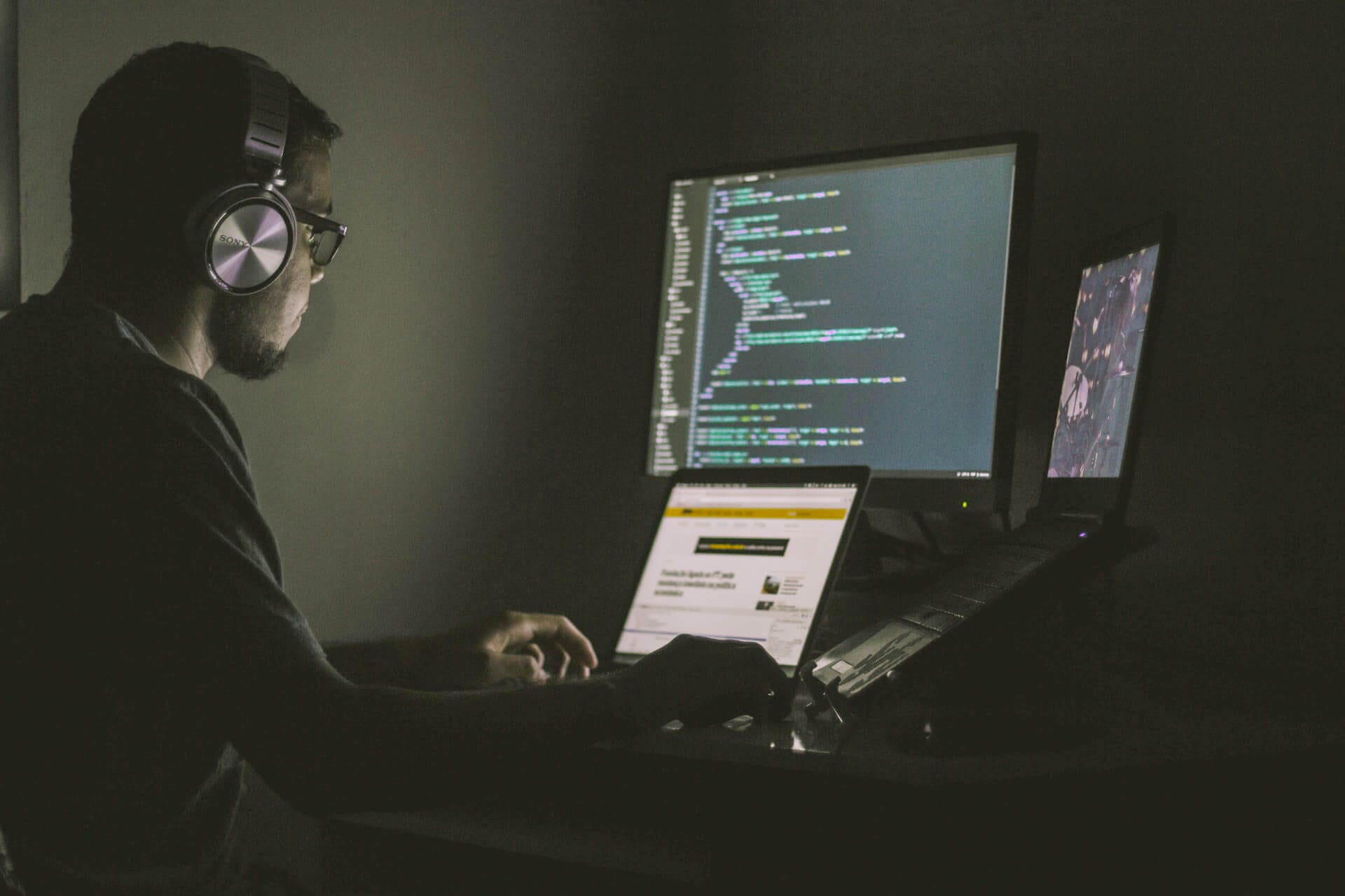 A developer wearing headphones, working in front of a screen and 2 laptops