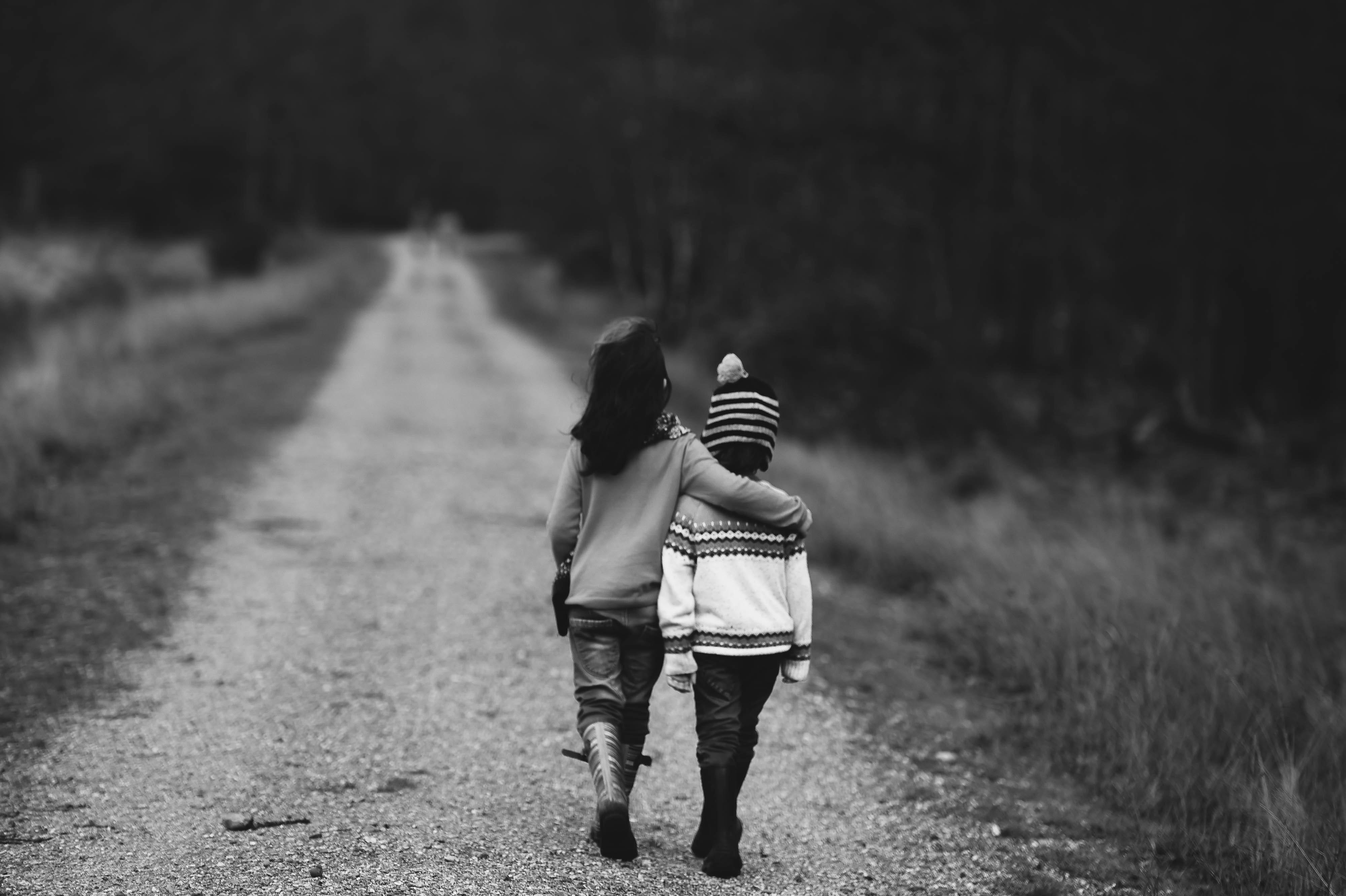 Rear view of 2 young children walking down a deserted dirt road. The taller has their arm around the smaller one’s shoulders.