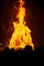 A large, orange bonfire reaches the top of the image before a black sky. At the bottom, heads on onlookers is cut off.