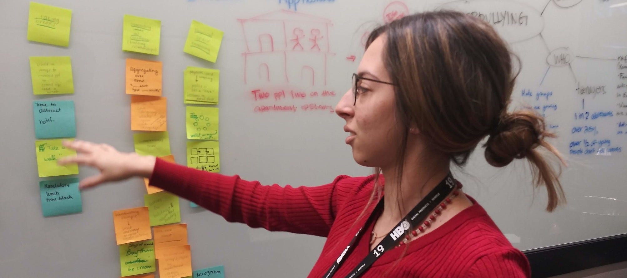 Woman with glasses in a red sweater pointing at Post-its on a whiteboard