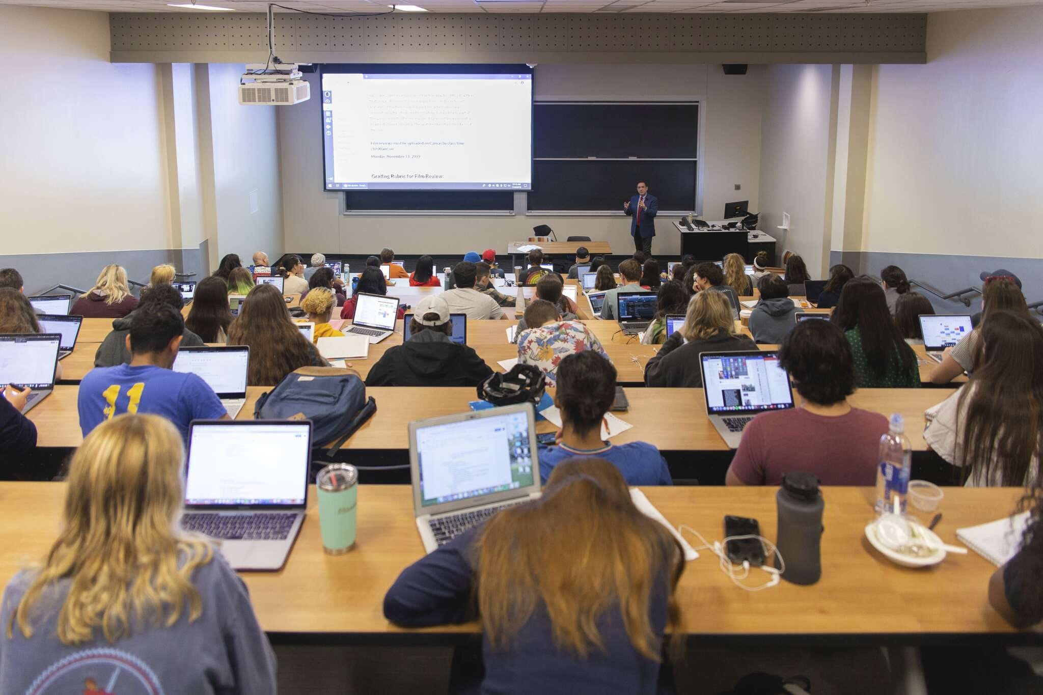Students fill every seat of a lecture hall. Thomas Garza stands in front of them next to a screen with information on it.