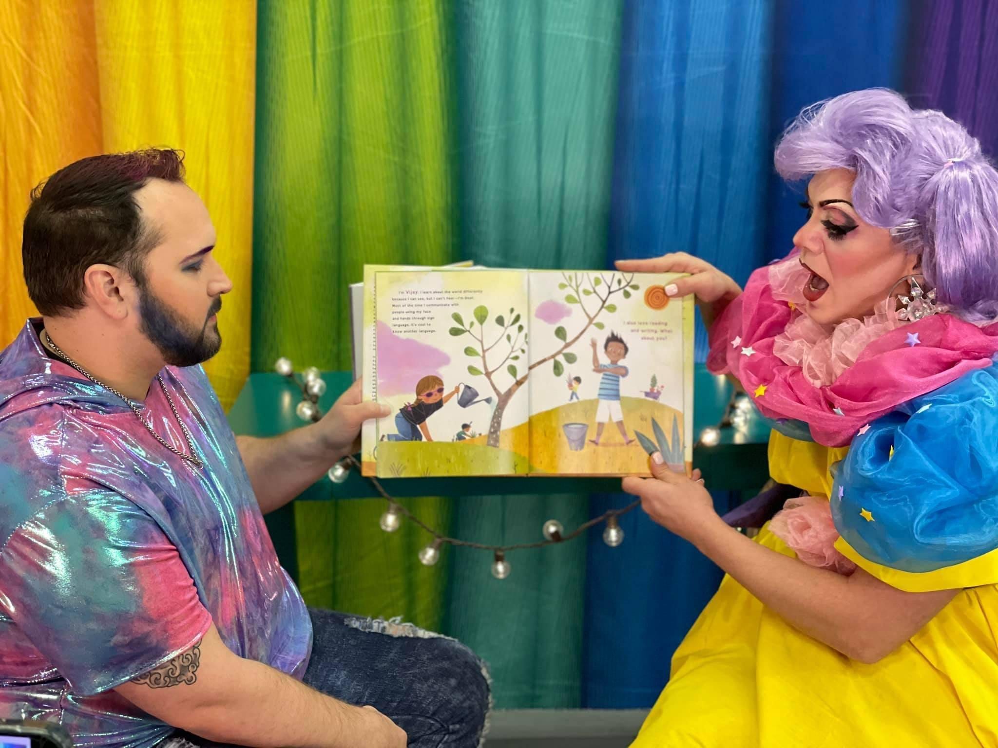 Two Drag Queen Story Time presenters are holding up a children’s book in front of a rainbow background as they read the story aloud