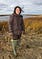 Woman in brown uniform and green rubber boots stands in grass with water behind.