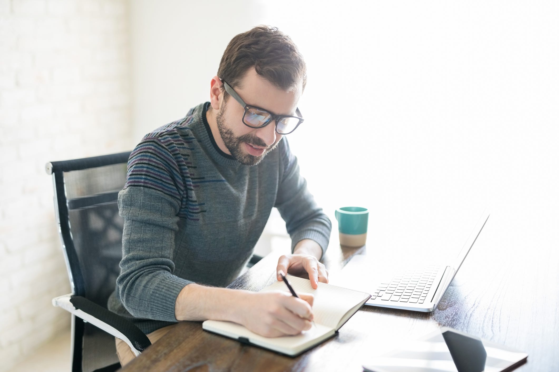 A man writes in his notebook while working at a desk with a laptop.