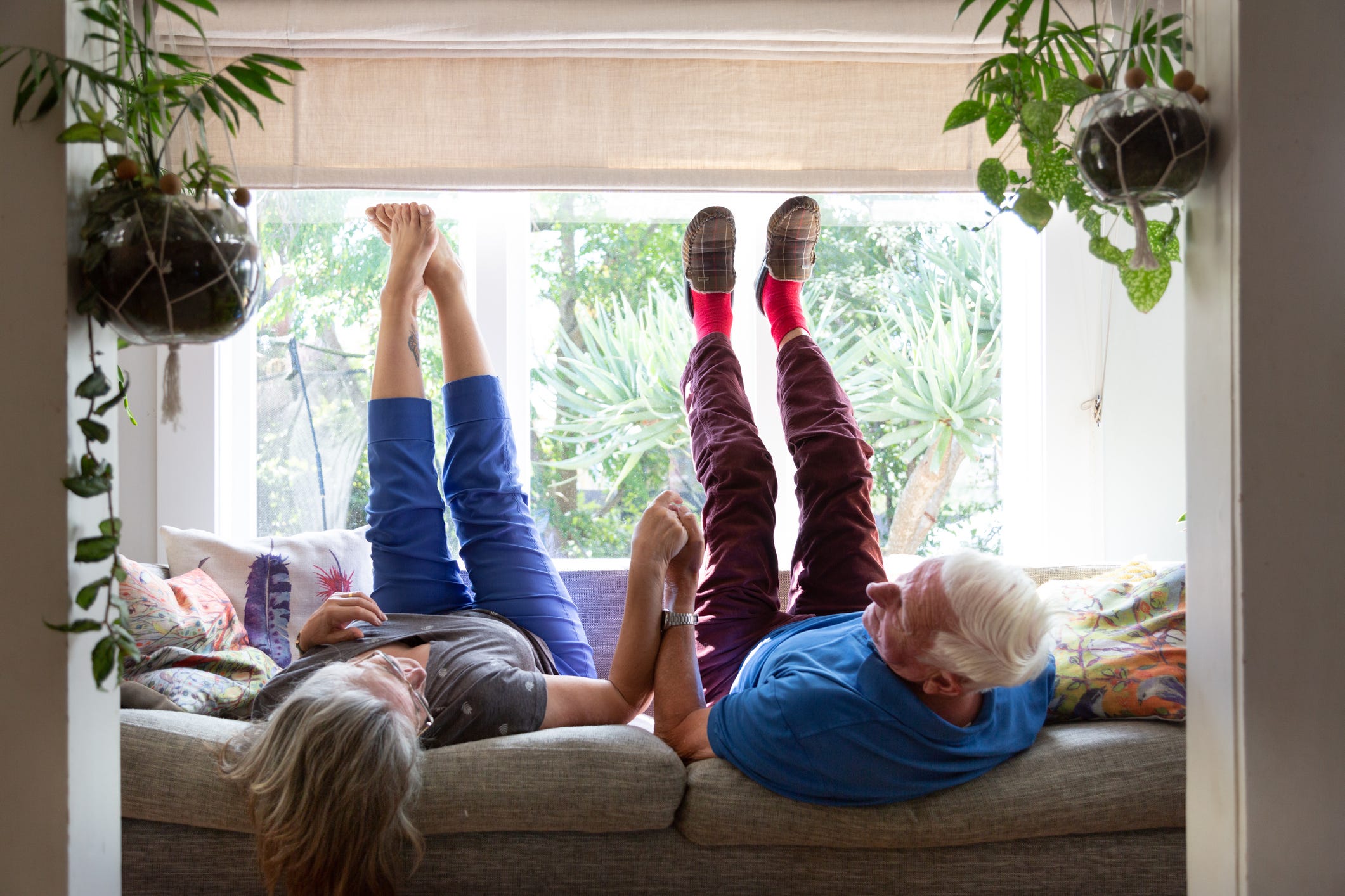 A senior couple lie next to each other on the sofa with their legs in the air. They are holding hands.
