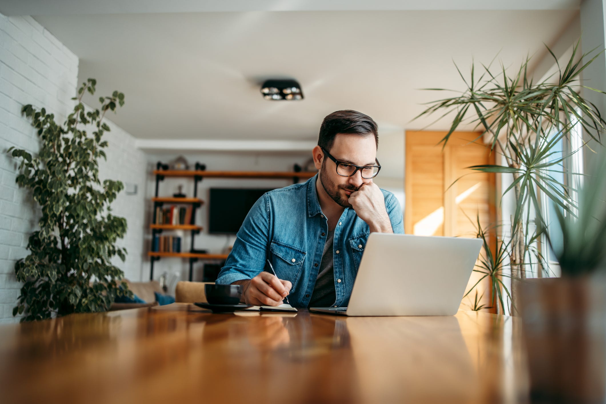 A photo of a man working on his computer at home as he is taking notes.