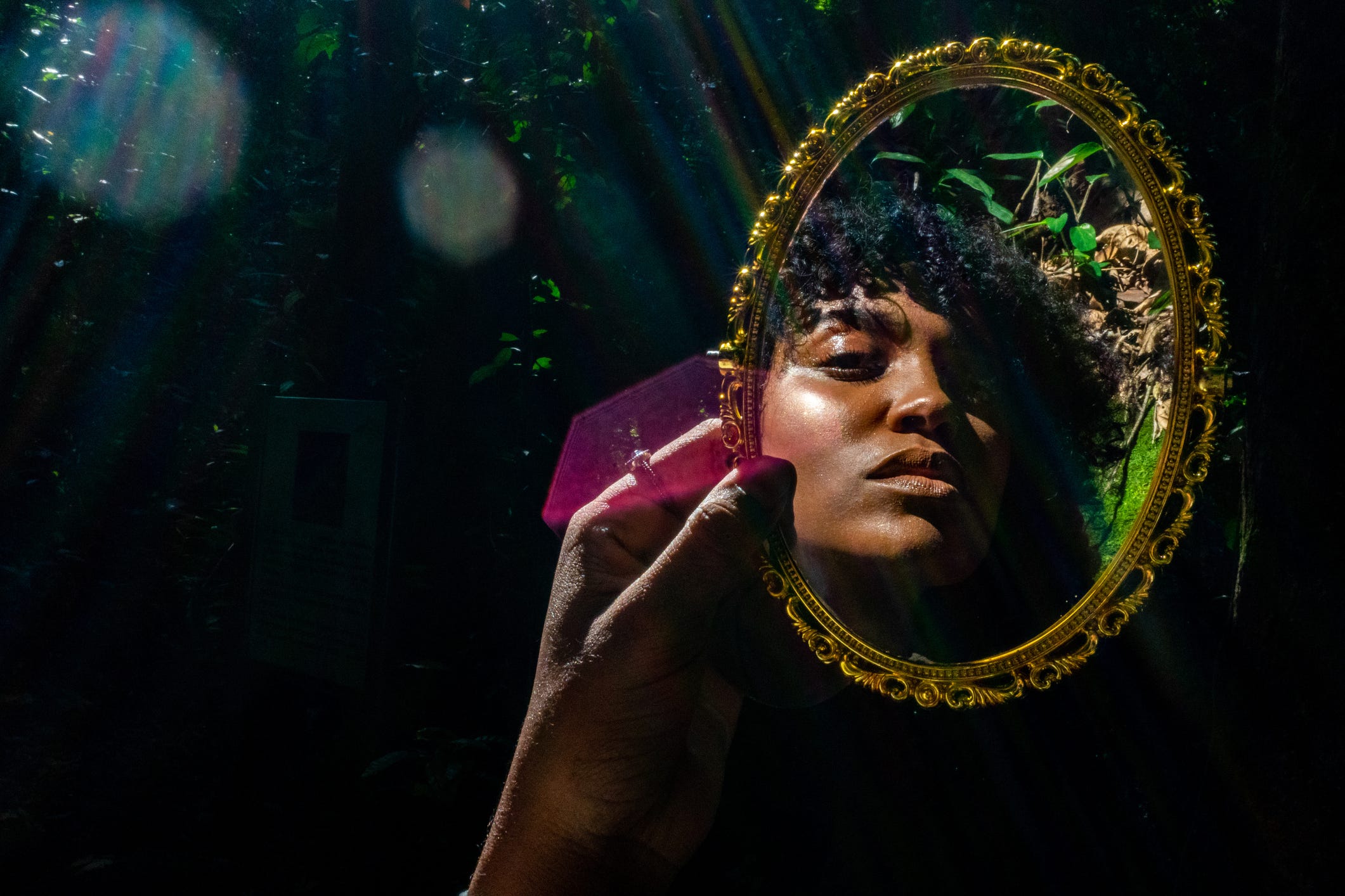 Young Black woman holding up and looking into a gilded mirror in the forest.