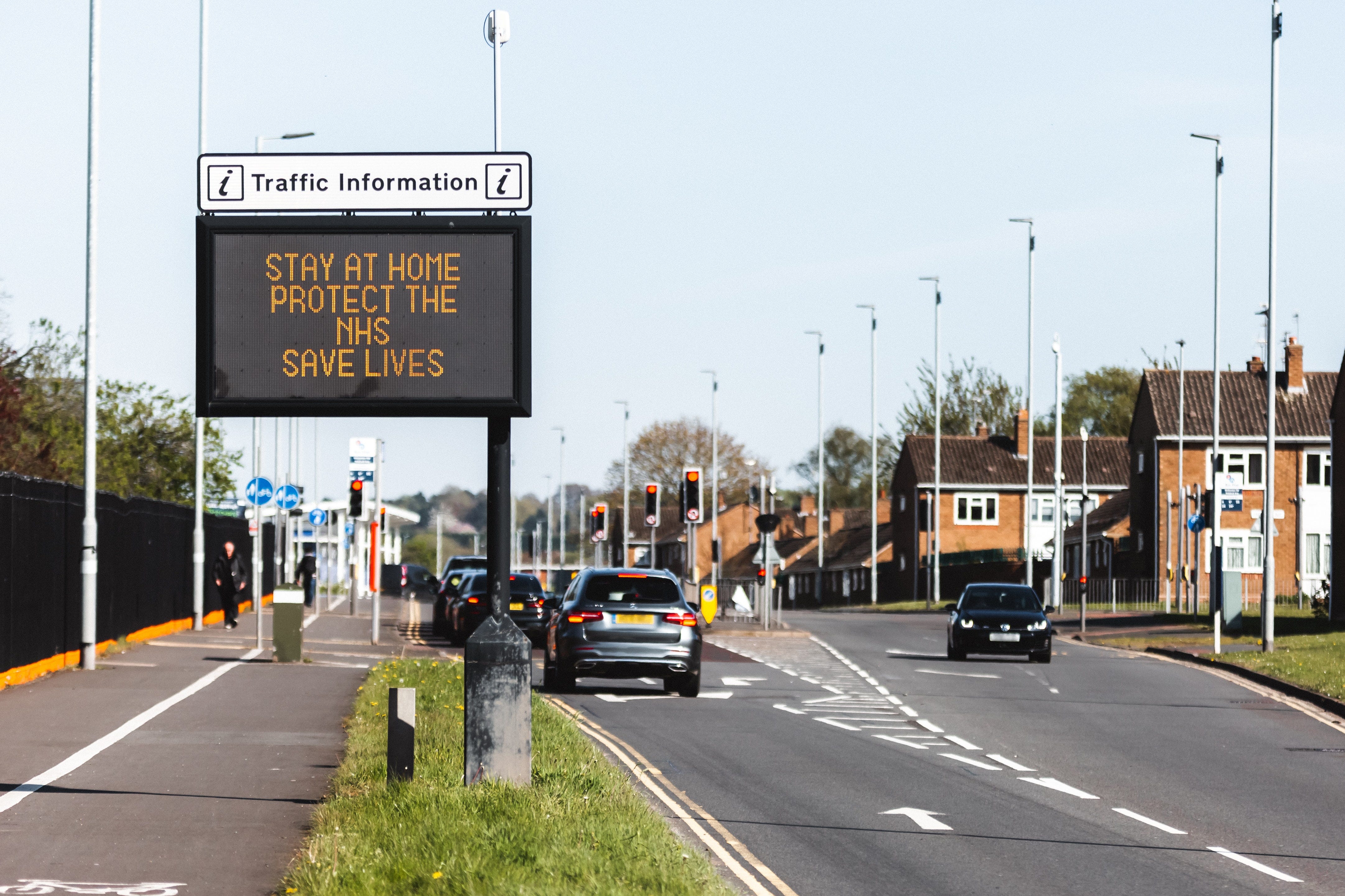 Vehicles on a road in the UK next to an electronic sign, where it reads: “Stay at Home. Protect the NHS. Save lives.”