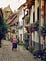 An elderly woman walking along a cobblestone street lined with houses showing off numerous flower boxes