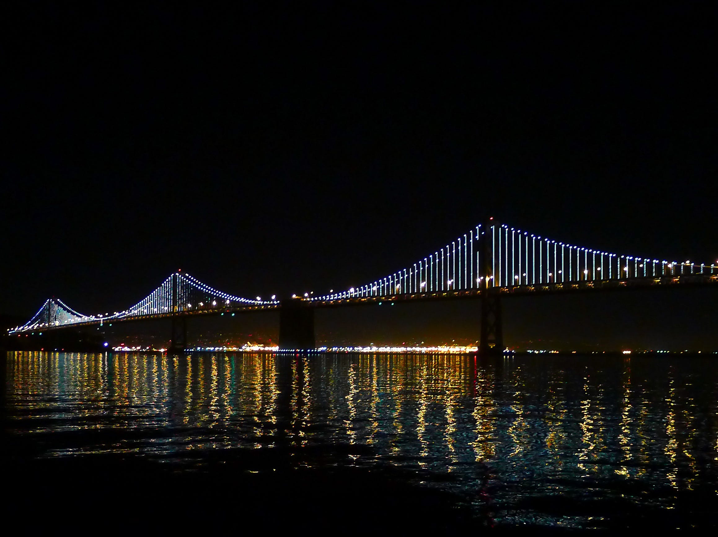 The Bay Bridge with its illuminated cables, as seen at night, above still, reflective waters.