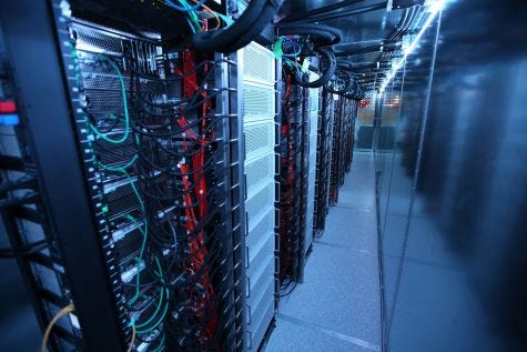 A narrow passageway with a network of computer racks on the left, and a metal wall on the right.