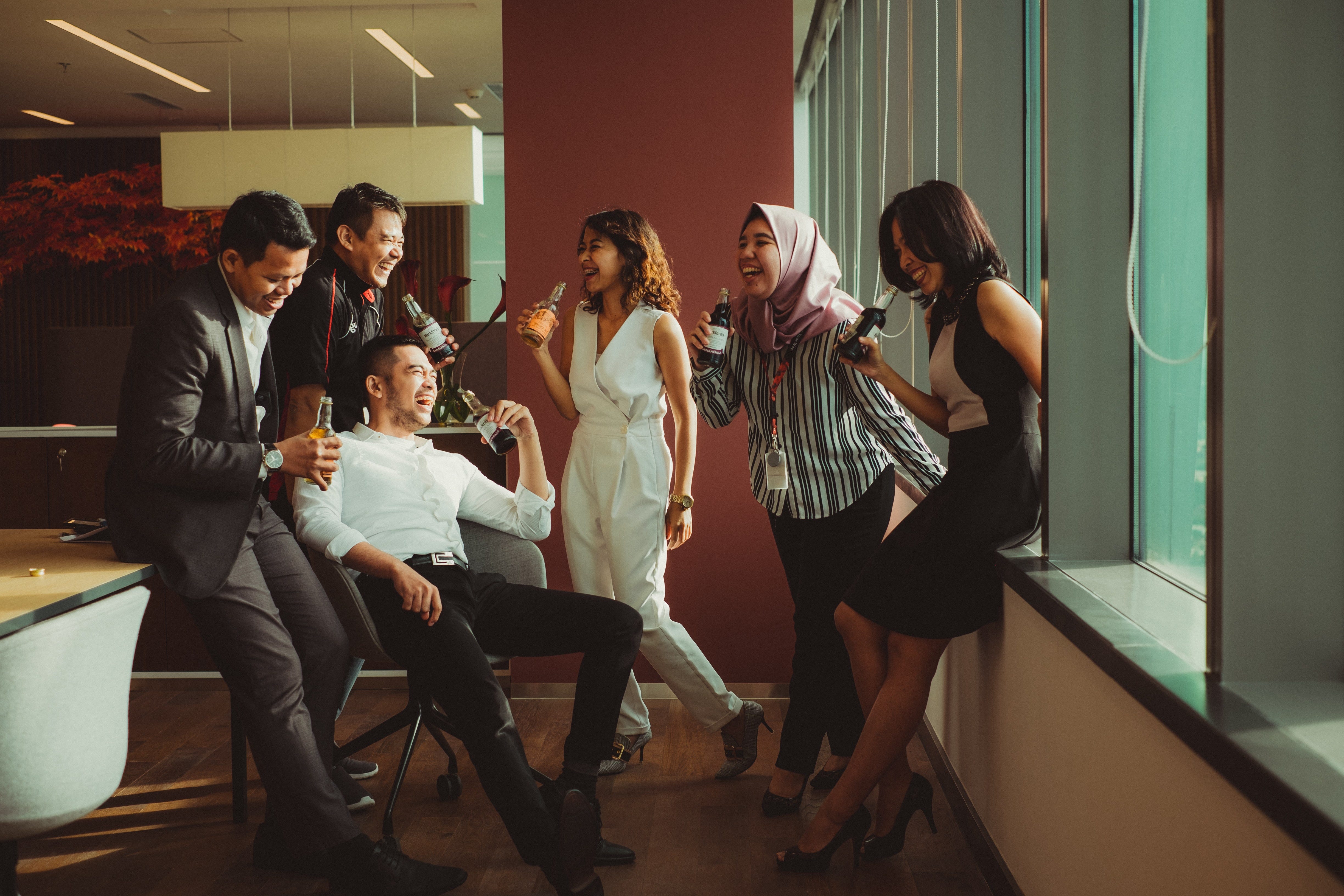 Seis personas de pie en la oficina, riendo y bebiendo cerveza