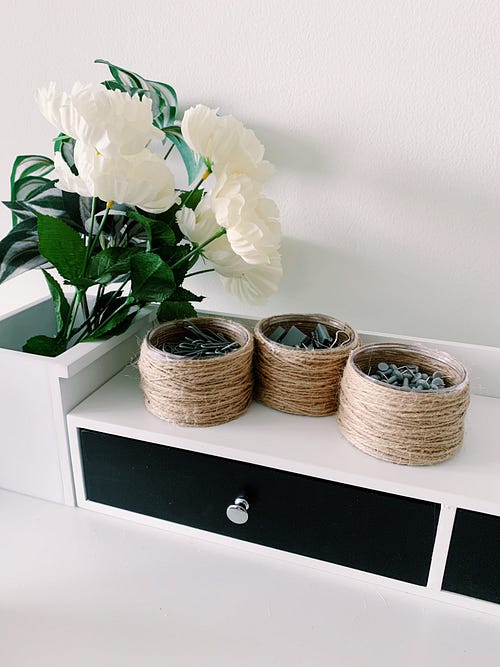 A white desk with twine wrapped plastic containers that were repurposed and to use for holding stationery items such as paper clips, push pins, and binder clips. There is also a little bouquet with white flowers and green leaves.