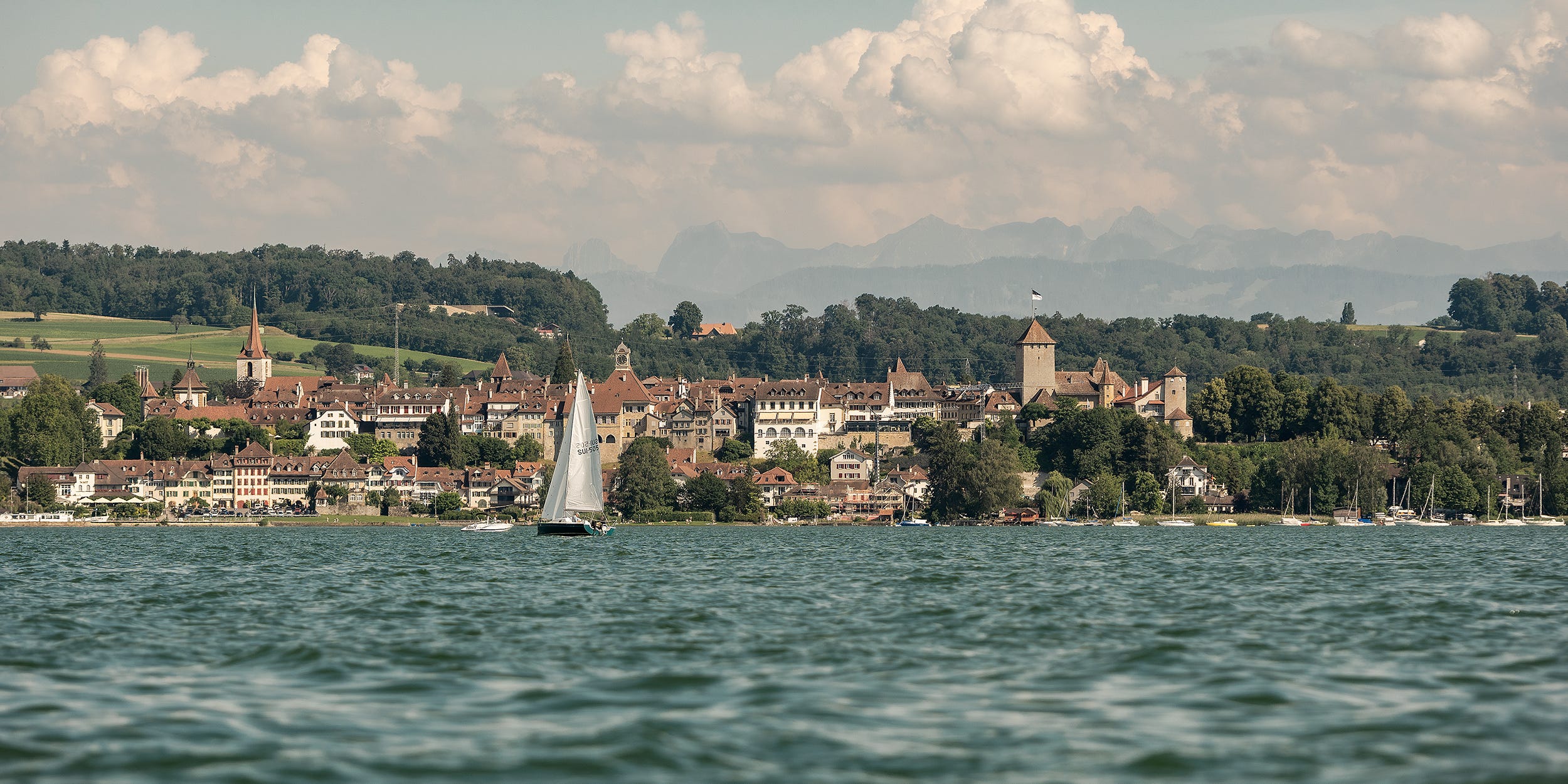 Blick vom See auf die Stadt Murten und die dahinter liegenden Hügelzüge.