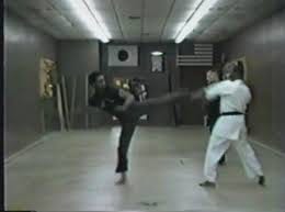 A low resolution frame showing a man in black sparring with someone in white, they are in a small martial arts dojo room.