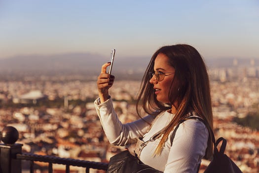 Photo Of Woman Using Mobile Phone. she’s set against a cityscape in the background. she’s probably taking a picture. she looks happy. i picked this photo because she’s on her phone, potentially making money, while out travelling.