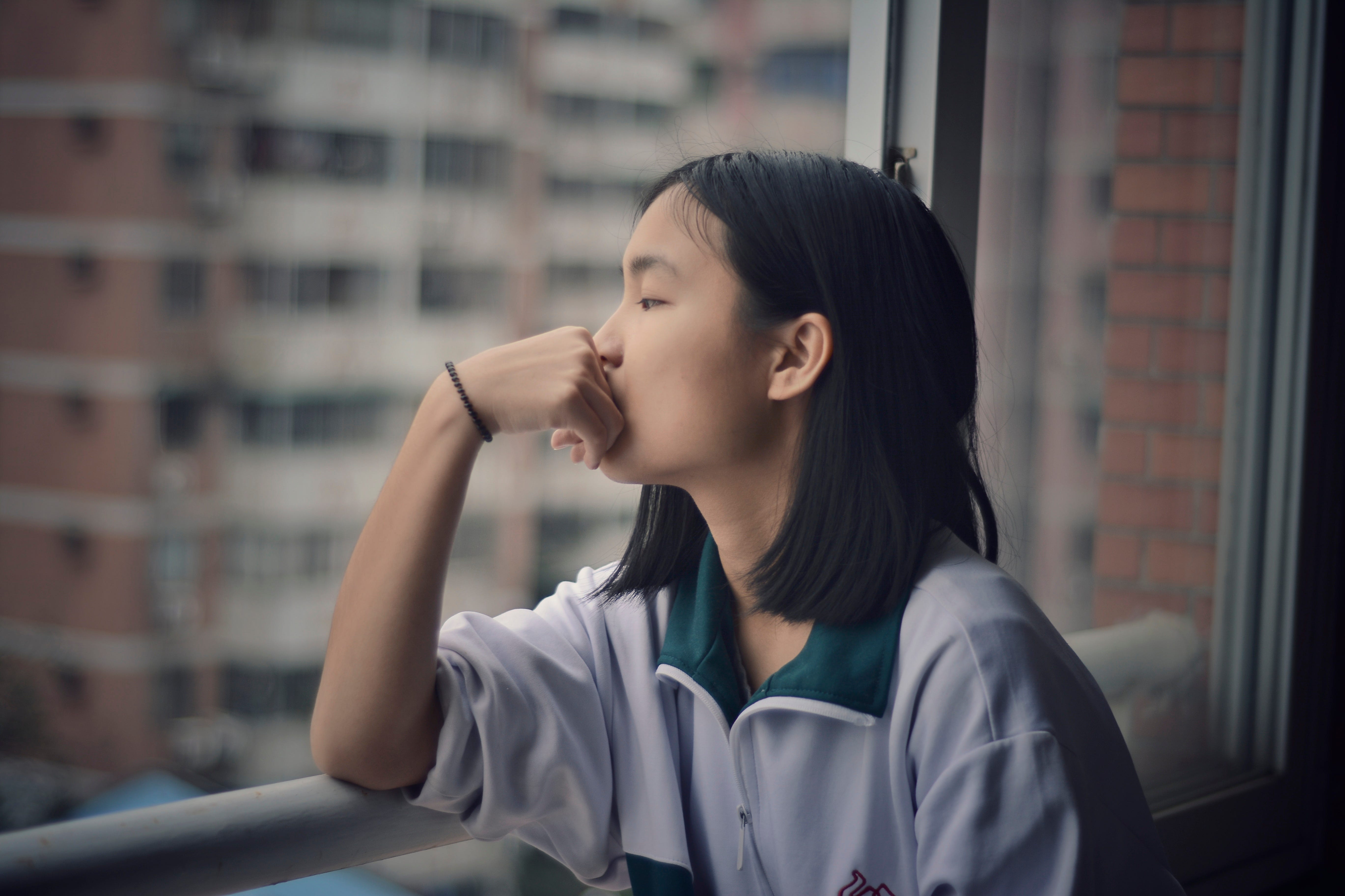 A young woman leans on a railing, with her hand next to her face, as she looks out the window in deep thought.