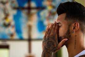 A young man, with tattooed hands covering his face, praying in a church.