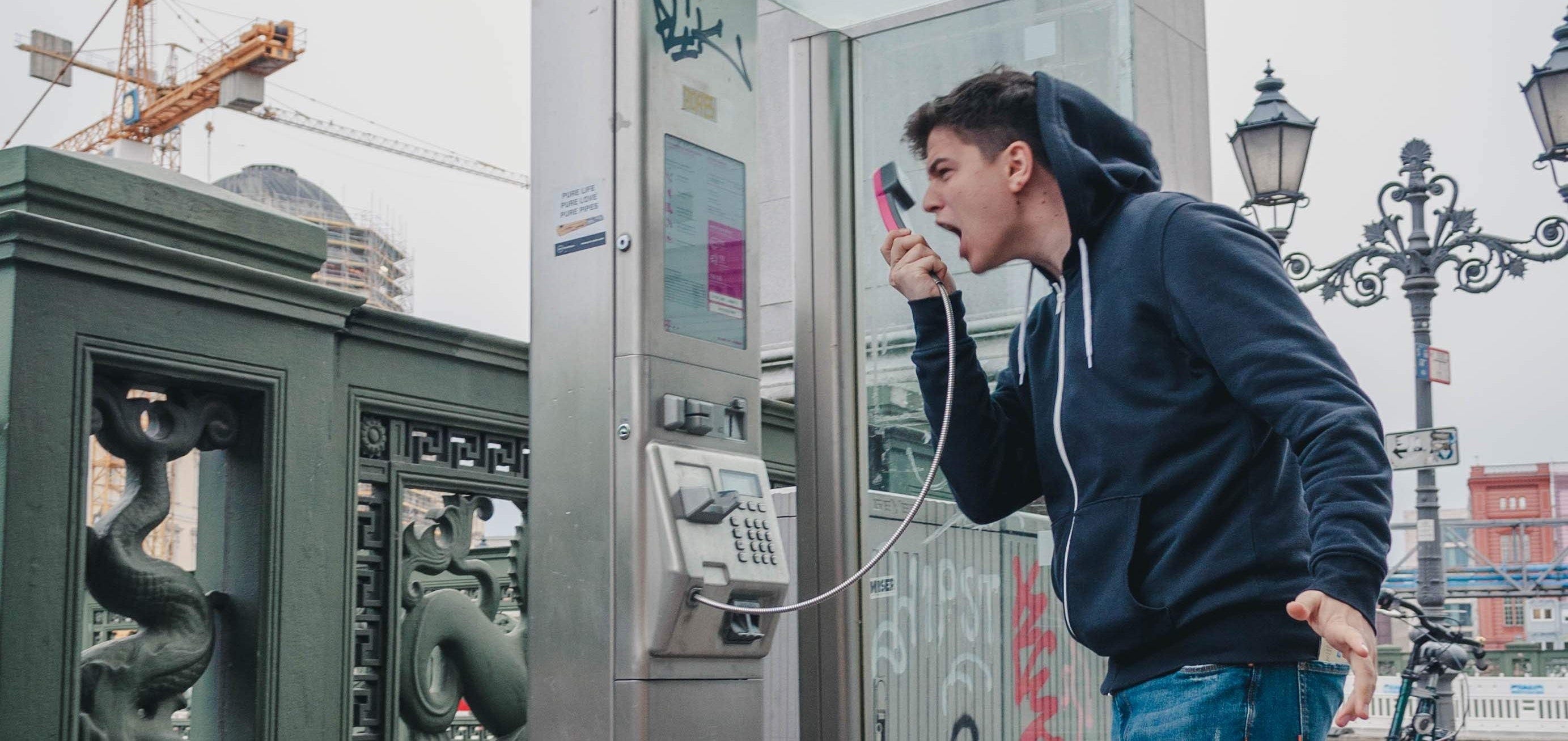This is an image of a white guy in a jeans and a dark blue hoodie who is screaming into the receiver of a payphone and gesturing angrily.