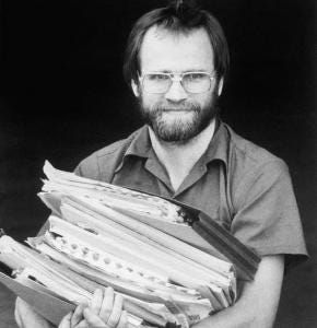 A bearded man in glasses carrying a large stack of bound papers
