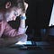 A man at his desk in the dark. He is in front of a computer while looking down at his iPad and holding an Apple Pencil.