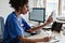A photo of a nurse sitting at a desk. She is doing a telehealth appointment, holding up a phone using the video function- whilst taking notes on a clipboard.