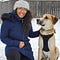 Catherine smiles and kneels next to Mickey (a beige and white medium-sized dog) in the snow