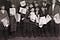 Newsboys holding up their papers for sale, dressed in black caps, jackets, knickers, socks, and boots.