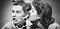 Black and white photo of one woman whispering into another woman’s ear, with the words “It’s a surprise!”