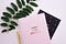 A pink journal rests on top of a black journal. A plant and golden pen are placed on the side.