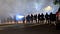 A line of police officers in riot gear standing in clouds of tear gas. They wear gas masks and carry batons and weapons