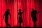 Three actors’ silhouettes in front of a red theatre curtain on a stage