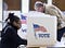 A black woman in a black coat with braids pulled back in a bun sits at a table with a cubicle decorated with vote and a flag