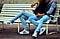 Couple sitting on wood slat bench wearing blue jeans and white tennis shoes