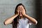 A person with long hair stands in front of a gray background, covering their eyes with their hands.